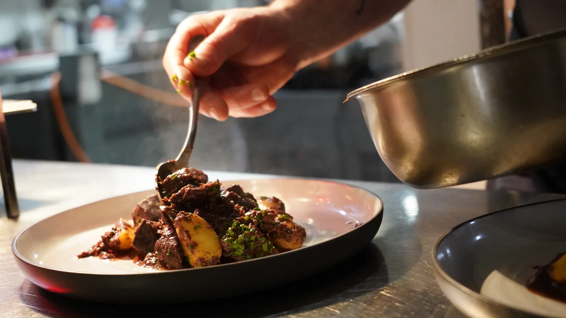 Un plat dressé en cuisine à l'Étoile Blanche, ragoût nappé à la cuillère, vapeur et lumière chaude